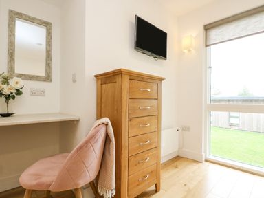 A wooden chest of drawers under a wall-mounted television next to a window and a pink chair with a blanket at Birch in Truro