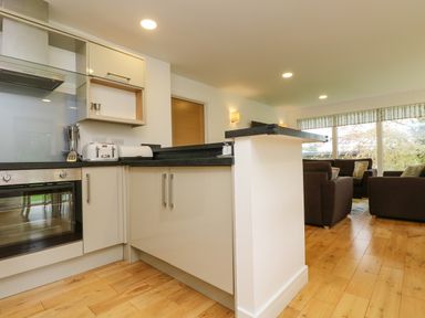 A kitchen with black countertops and beige cabinets next to a living room with brown sofas and large windows at Elm in Truro