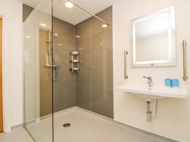 A bathroom with a walk in shower with glass partition a wall mounted sink with two blue cups and a square illuminated mirror at Elm in Truro