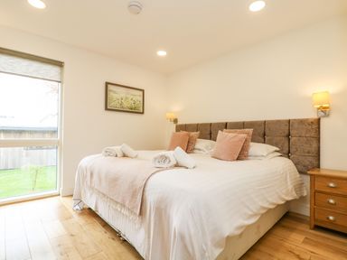 A bedroom with a large bed with pillows and rolled towels a window with a view of a lawn a bedside table with drawers and wall lamps Elm Truro