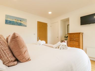 A bedroom with a bed with three pillows and rolled towels a wooden dresser a wall-mounted TV and a wooden door at Elm in Truro