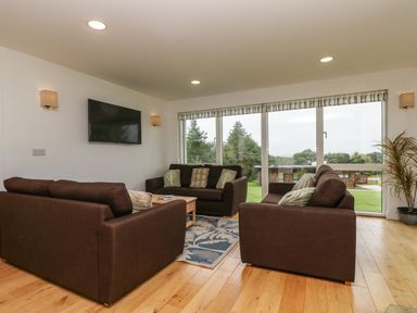 A living room with three brown sofas a wall-mounted TV and large windows showing an outdoor view at Hazel in Truro