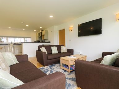 A living room with brown sofas, a wooden coffee table, a wall-mounted television, and a kitchen area in the background at Hazel in Truro