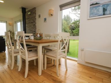 A dining area with a wooden table and six white chairs next to a large window showing a green lawn at Hazel in Truro