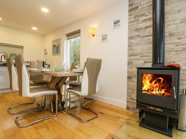 A dining area with a wooden table and beige chairs next to a black wood-burning stove with a fire at Juniper in Truro