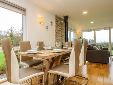 A dining area with a wooden table set with plates and glasses and leather chairs next to a living room with sofas and large windows at Juniper in Truro
