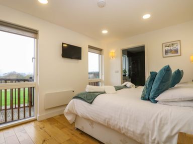 A bedroom with a bed arranged with rolled towels and blue pillows a wall-mounted TV two windows and a door leading to a bathroom at Juniper in Truro