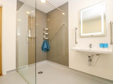 A bathroom with a walk-in shower with glass partition a sink with two blue cups and an illuminated mirror at Juniper in Truro