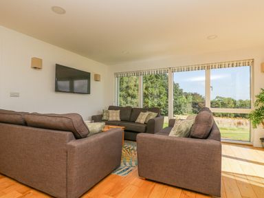 A living room with three gray sofas a coffee table a wall mounted TV and large windows showing trees outside at Rowan in Truro