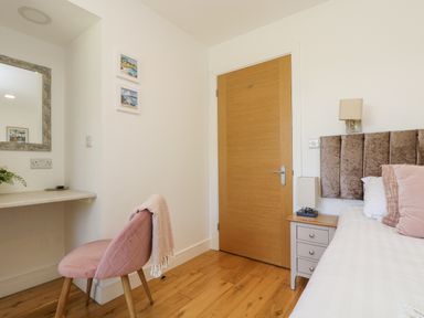 A bedroom corner with a wooden door a bedside table with a lamp part of a bed with pillows a pink chair with a throw next to a wall-mounted desk and mirror at Rowan in Truro