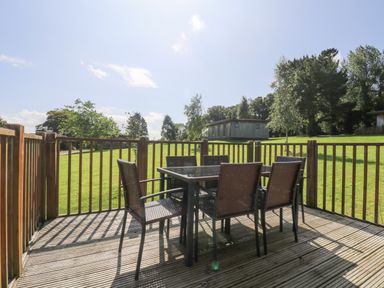 A wooden deck with a glass top table and six wicker chairs overlooking a grassy lawn with trees at Rowan in Truro