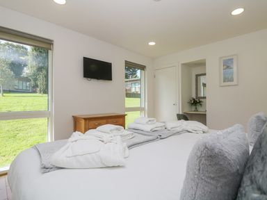 A bedroom with a bed, folded towels on the bed, a wooden dresser, a wall-mounted TV, two windows showing grass outside, and a small desk with a mirror and flowers at Willow in Truro