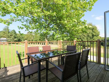 An outdoor wooden deck with a glass top table and six wicker chairs surrounded by trees and grass at Willow in Truro