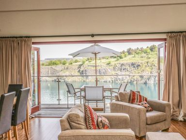 A living area with armchairs and dining chairs facing a balcony with table and umbrella overlooking a lake and rocky hillside at Boavista in Carnforth