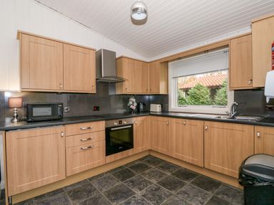 A kitchen with wooden cabinets a black countertop a microwave oven a stove a sink and a window overlooking trees at Sunset Lodge in Tattershall