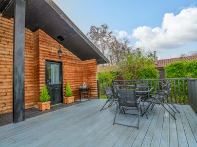 An outdoor wooden deck with a glass table and chairs beside a log cabin at Sunset Lodge in Tattershall