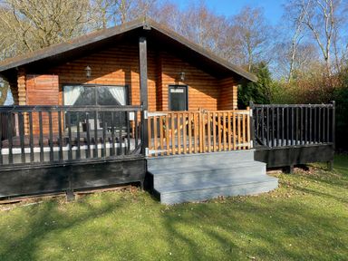 A wooden cabin with a porch and railing surrounded by trees and grass at Sunset Lodge in Tattershall
