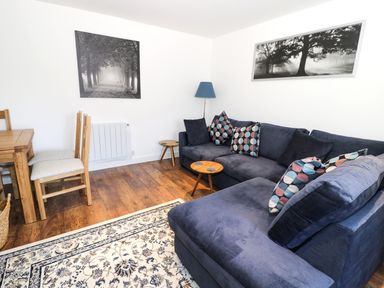 A living room with a blue sectional sofa with patterned cushions two wooden side tables a wooden dining table with chairs a floor lamp and black and white nature pictures on the walls at Skovhaven in Catton