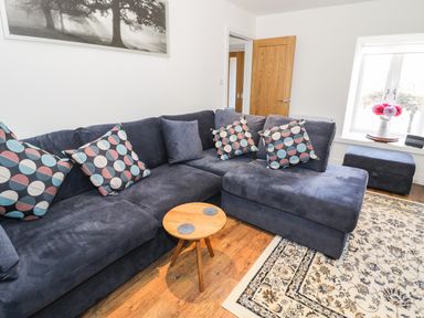 A living room with a dark blue sectional sofa with patterned cushions a small wooden table a window with flowers on the sill and a patterned rug on the floor at Skovhaven in Catton