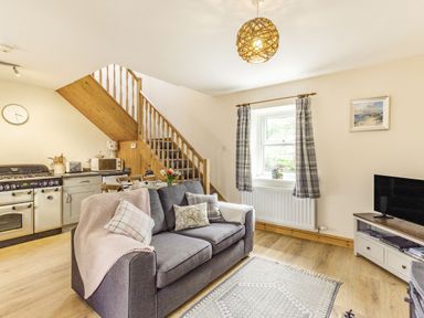 An open living room and kitchen with gray sofa dining table wooden stairs and television at Briar Cottage in Edlingham