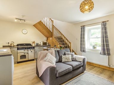 A kitchen and living area with a gray sofa under stairs next to a window with checked curtains at Briar Cottage in Edlingham