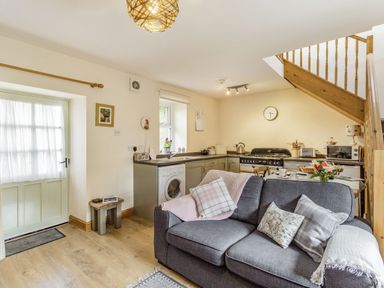 An open living room and kitchen area with a gray sofa pillows a washing machine and wooden stairs at Briar Cottage in Edlingham