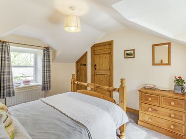 A bedroom with a wooden bed, dressers, two wooden doors, window with curtains, and a ceiling light at Briar Cottage in Edlingham