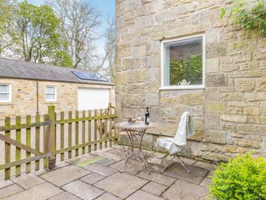An outdoor stone patio with a small round table and two metal chairs next to a stone wall with a window and a wooden gate at Briar Cottage in Edlingham