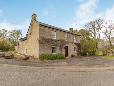An exterior view of a stone house with two chimneys and a red door by a paved driveway with rocks and trees at Briar Cottage in Edlingham