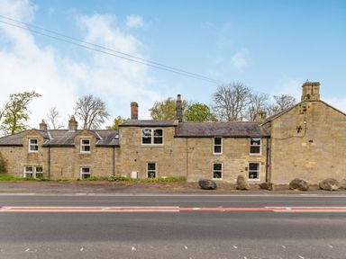 A stone building with multiple windows alongside a road with rocks in front at Briar Cottage in Edlingham