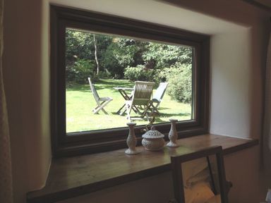 A window with ornaments on the wooden sill overlooking a garden with chairs and a table at Water Barn in Manaton near Lustleigh