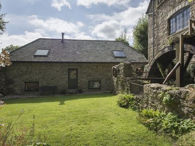 A stone building with windows skylights and a water wheel next to a lawn at Water Barn in Manaton near Lustleigh