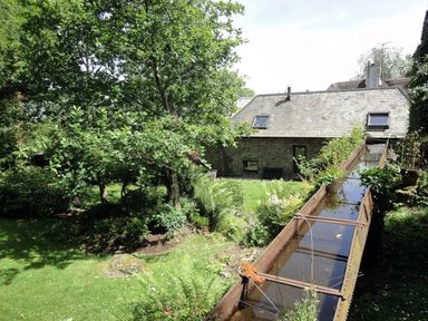 A garden with trees and plants next to a stone house with a water channel in the foreground at Water Barn in Manaton near Lustleigh