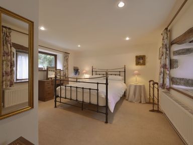 A bedroom with a metal bed frame white bedding wooden dresser with mirror table lamp and floral curtains at Water Barn in Manaton near Lustleigh