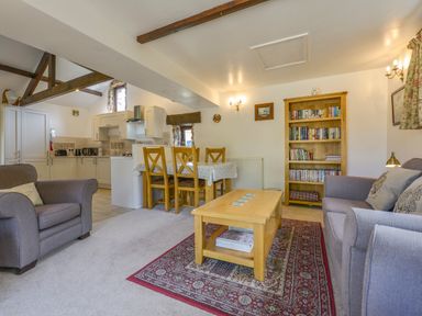 A living room with two armchairs a sofa a wooden coffee table with magazines a bookshelf and a dining table with chairs at Buster Barn in Sampford Spiney near Tavistock