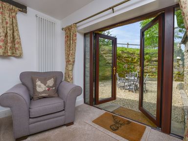 A living area with a gray armchair and a cushion by open wooden patio doors leading to an outdoor patio with a metal table and chairs at Buster Barn in Sampford Spiney near Tavistock