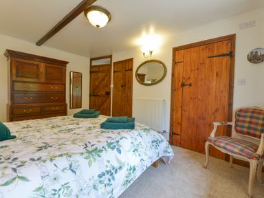 A bedroom with a bed covered by floral bedding two green towels wooden dresser doors oval mirror and a checkered armchair at Buster Barn in Sampford Spiney near Tavistock