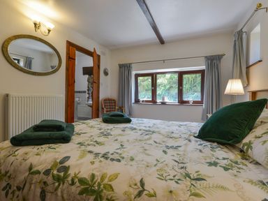 A bedroom with a bed with floral bedding green pillows and towels next to a window with curtains a chair beside an open door and a mirror on the wall at Buster Barn in Sampford Spiney near Tavistock