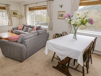 A dining table with a white tablecloth and a vase of flowers next to a living area with gray sofas in a room at The Dene in Betws-y-Coed