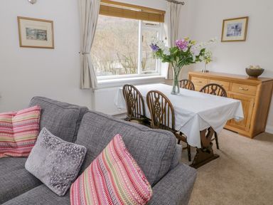 A dining area with a table covered by a white cloth and a vase of flowers surrounded by wooden chairs next to a grey sofa with colorful cushions and a wooden sideboard with framed pictures on the walls at The Dene in Betws-y-Coed