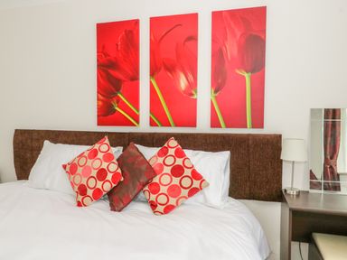 A bed with red patterned cushions and a brown headboard below three red flower paintings at The Dene in Betws-y-Coed