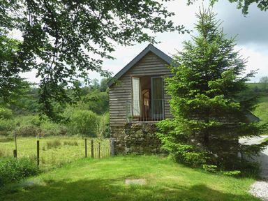 A small wooden house with a stone foundation surrounded by trees and grass at The Linhay in Lydford