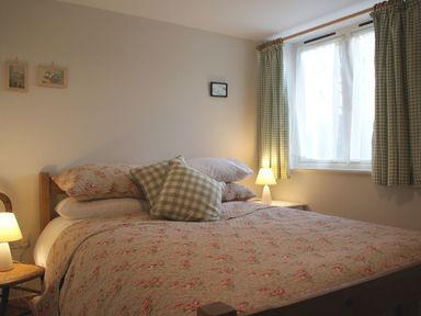 A bedroom with a bed covered with floral bedspread and checkered pillows next to two bedside lamps and a window with checkered curtains at The Stone Barn Cottage in Holne