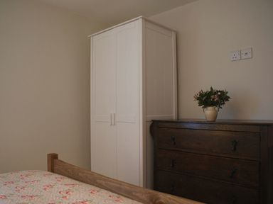 A bedroom corner with a white wardrobe a wooden dresser with a vase of flowers on top and a bed with floral bedding at The Stone Barn Cottage in Holne