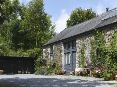 Stone cottage with gray door and windows surrounded by plants and flowers with gravel driveway at The Stone Barn Cottage in Holne