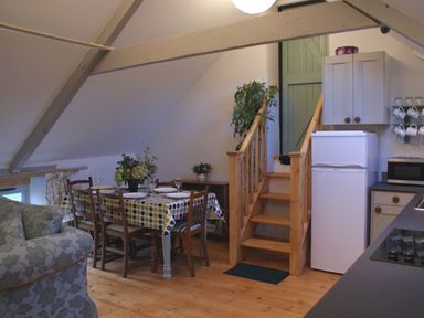 A dining area with a wooden table and chairs next to wooden stairs and a kitchen with a refrigerator at The Stone Barn Cottage in Holne