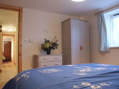 A bedroom with a blue patterned bedspread a white chest of drawers with flowers and a wardrobe near a window with checked curtains at The Stone Barn Cottage in Holne