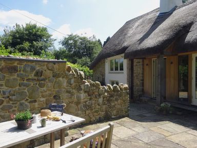 An outdoor stone patio with a wooden table and bench next to a stone wall and a cottage with a thatched roof at Cullaford Cottage in Scorriton near Ashburton