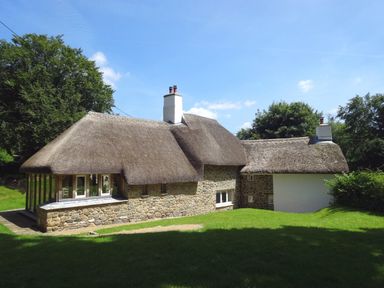 A stone cottage with thatched roof and chimneys surrounded by grass and trees at Cullaford Cottage in Scorriton near Ashburton