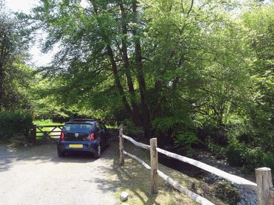 A black car parked on a gravel road near a wooden fence and large green trees at Cullaford Cottage in Scorriton near Ashburton